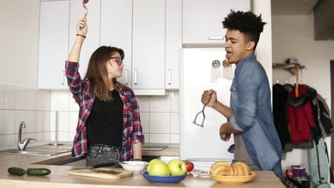 pareja linda haciendo tonterías, bailando en la cocina con utensilios de cocina. chica caucásica atractiva y su novio mulato vistiendo elegantes camisas hipster, disfrutando de su tiempo juntos.