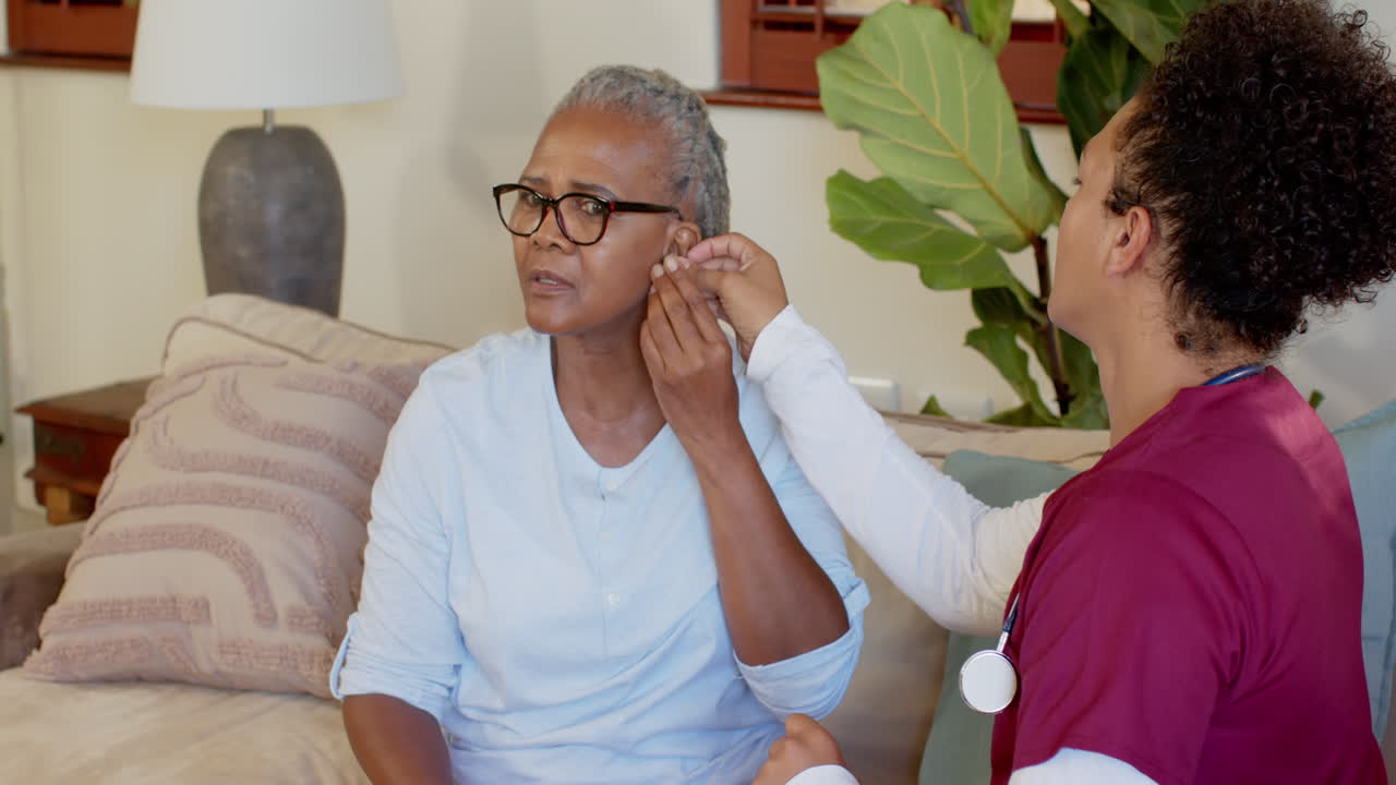 Senior woman adjusting hearing aid while caregiver in scrubs offers assistance, at home