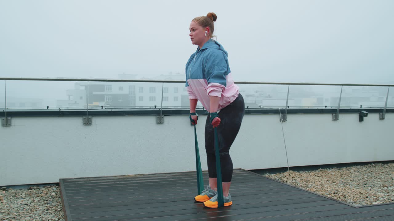 스포츠 의류 를 입은 스포츠 소녀 는 야외 에 있는 집 지붕 에 고무 밴드 를 사용 하여 요가 스트레칭 운동 을 한다