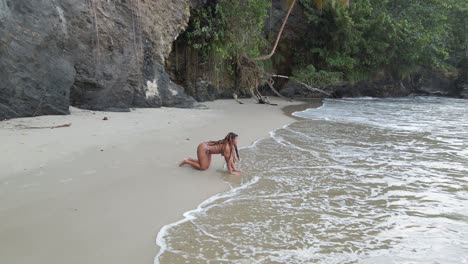 Aerial-view-of-a-girl-in-a-bikini-playing-at-the-shoreline-with-rocky-cliffs-in-the-background