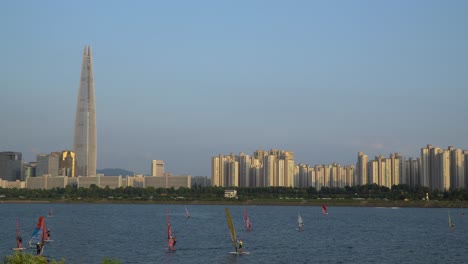 Grupo-De-Windsurfistas-Irreconocibles-Surfeando-En-El-Río-Han,-Seúl-Al-Atardecer,-Famoso-Edificio-Emblemático-De-La-Torre-Lotter-En-El-Fondo