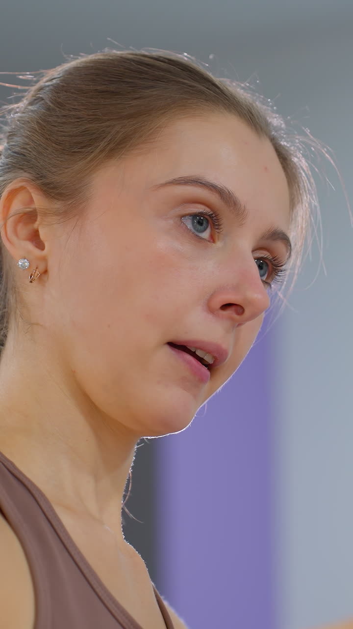 mujer con ojos azules y cabello atado hacia atrás se centra mientras ayuda con un estiramiento del brazo, el fondo presenta un entorno de gimnasio borroso con iluminación suave