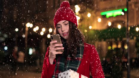 vista cercana de una mujer caucásica con abrigo rojo sosteniendo un regalo y usando un teléfono inteligente en la calle mientras nieva en navidad