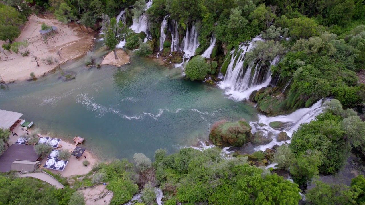 Cascata di Kravica che sfocia in una calma piscina turchese delimitata da alberi e sponde rocciose in Bosnia, approccio aereo con tilt down