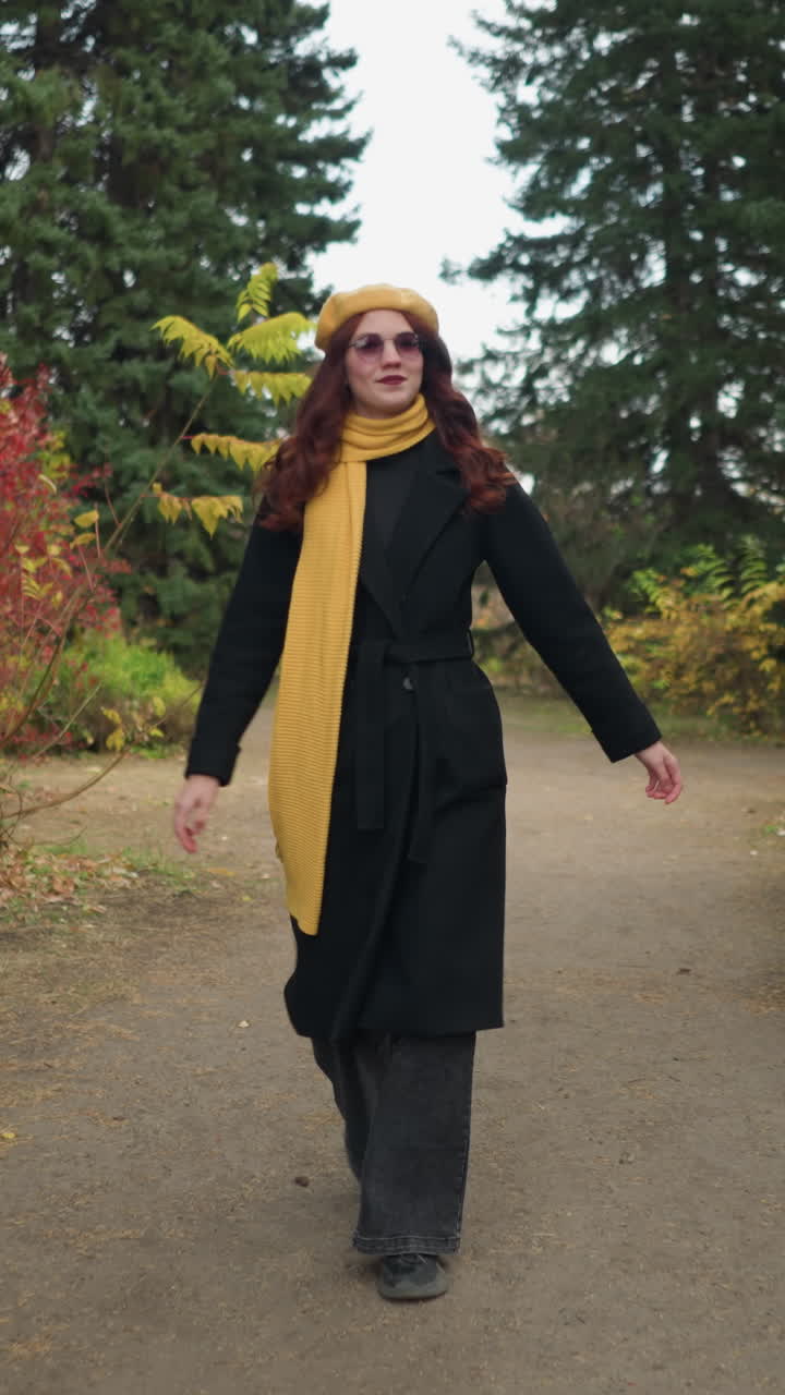 Turista con occhiali da sole e lunghi capelli rossi che indossa un basco giallo, una sciarpa abbinata e un cappotto nero, che cammina e sorride mentre si guarda intorno in un giardino vivace con un colorato fogliame autunnale.