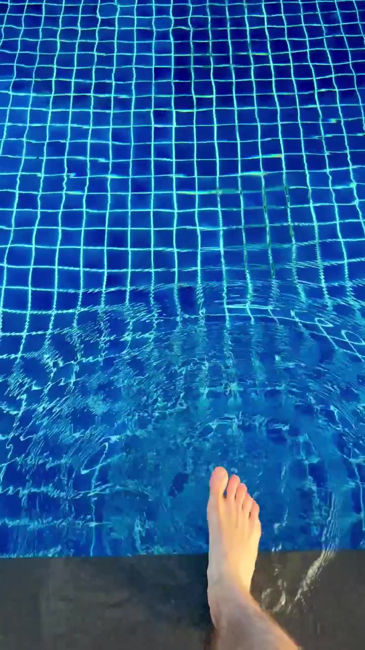 Seorang pria mencelupkan kaki telanjang ke dalam kolam renang biru. Menguji suhu air sebelum masuk. Rekaman POV vertikal yang memperlihatkan riak menyebar di permukaan dan menyampaikan rasa antisipasi serta keraguan