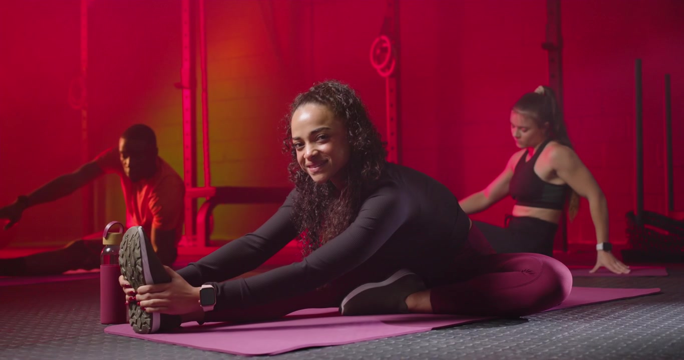 Diverse gym group during warmup stretching on mats, center woman holding shoe on bolster, smiling