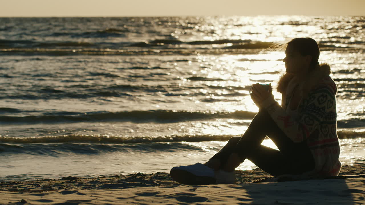 시원한 바람이 부는 날 해변에 앉아 있는 젊은 여성은 컵에서 뜨거운 음료를 마시고 공동에서 따뜻하게 유지