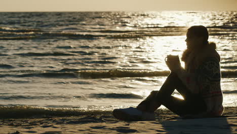 Junge-Frau,-Die-An-Einem-Kühlen,-Windigen-Tag-Am-Strand-Sitzt-Und-Ein-Heißes-Getränk-Aus-Einer-Tasse-Trinkt,-Hält-Sich-In-Co-Warm