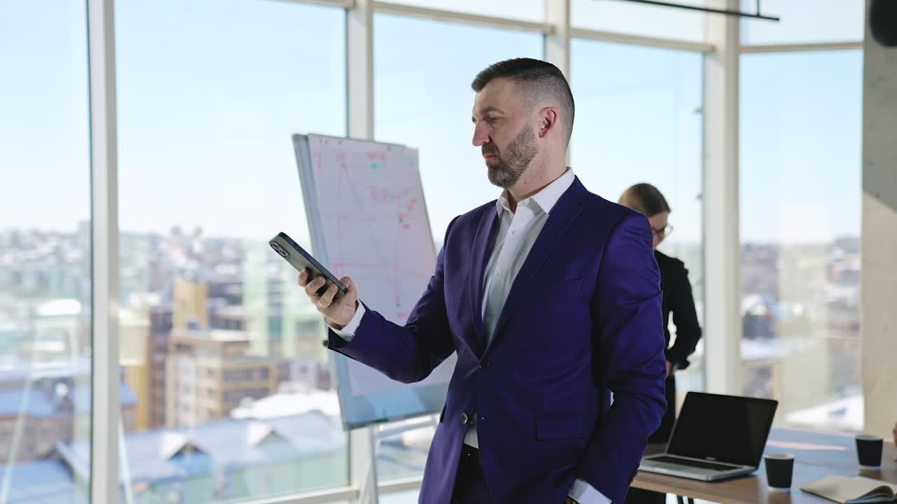 Working people busy in their big light office. Bearded businessman calling someone on the phone. Employees at backdrop stand at the chart. Blurred backdrop.