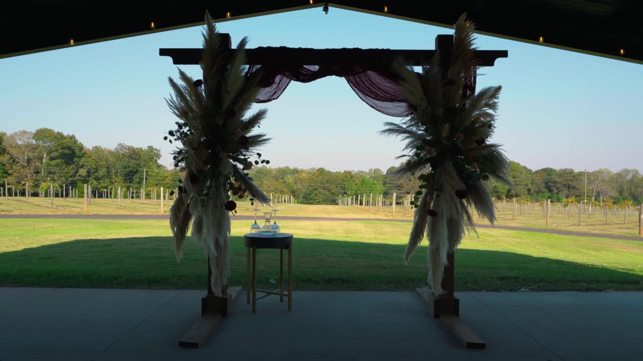 A straight-on slow push-in shot of a beautifully decorated wedding arch featuring pampas grass and florals, with a vineyard backdrop in South Georgia.
