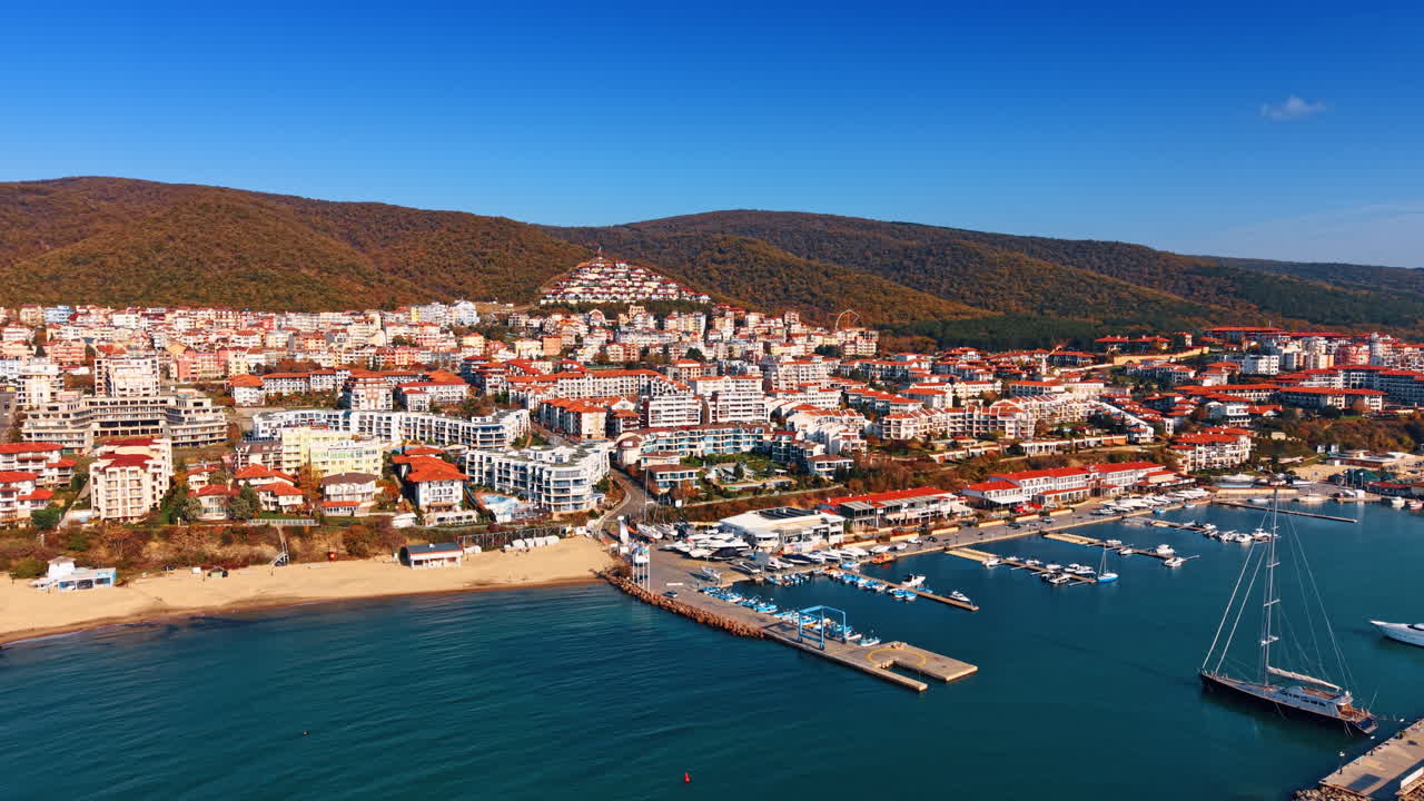 Vista aerea di una città costiera con porto turistico e montagne. Panorama di una località marittima con tetti in terracotta, un porto turistico e colline boscose sotto un cielo azzurro.