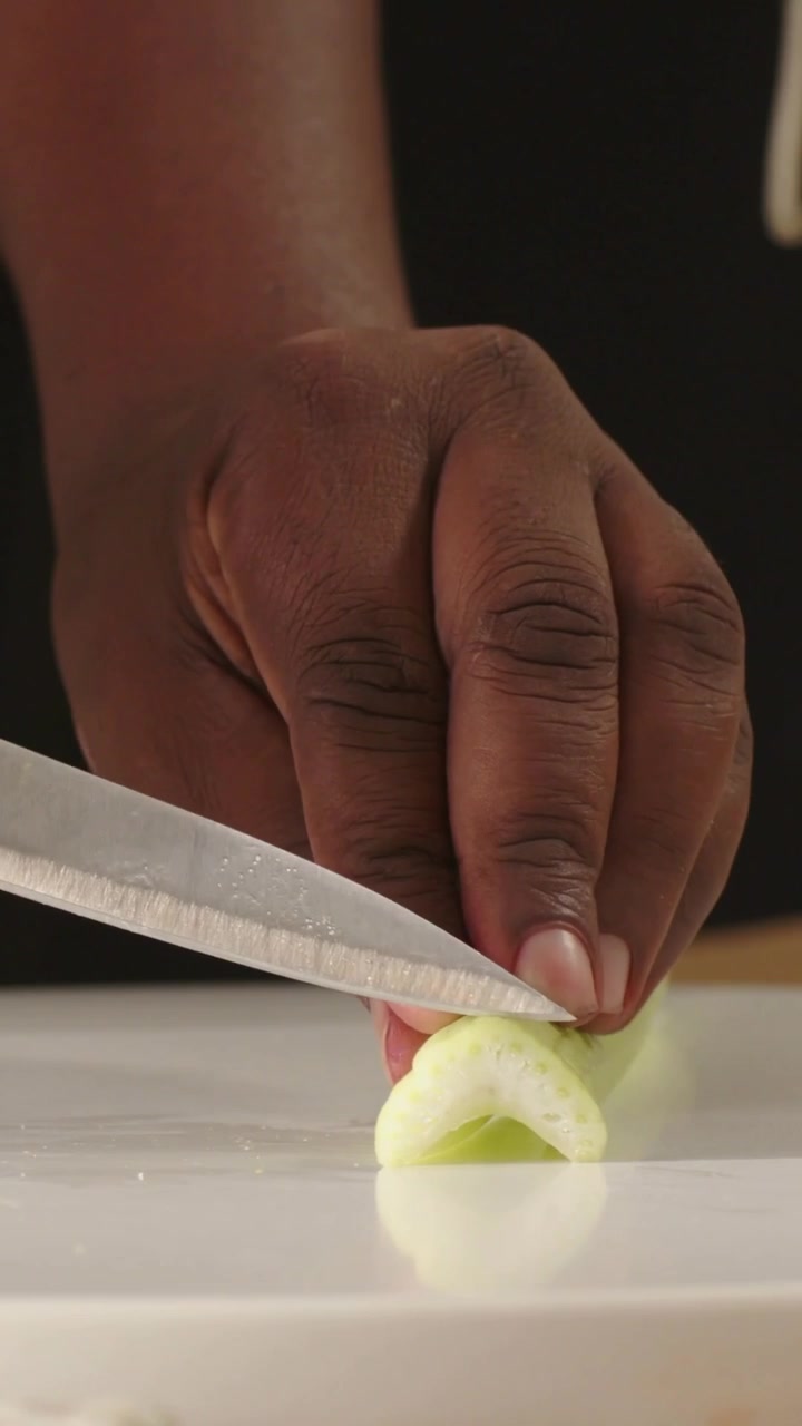 Video vertical: Manos con blusa oscura rebanando apio en una tabla sobre la encimera, cuchillo de chef entrando