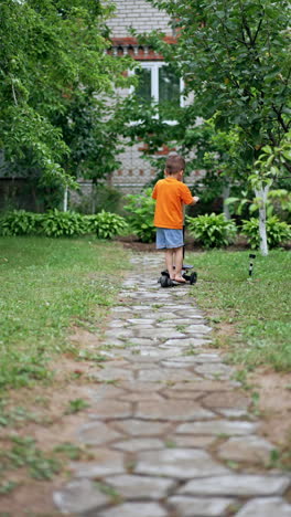 rear view of a baby boy in orange t-shirt and shorts riding a scooter. active kid having fun in the backyard in summer. vertical video.
