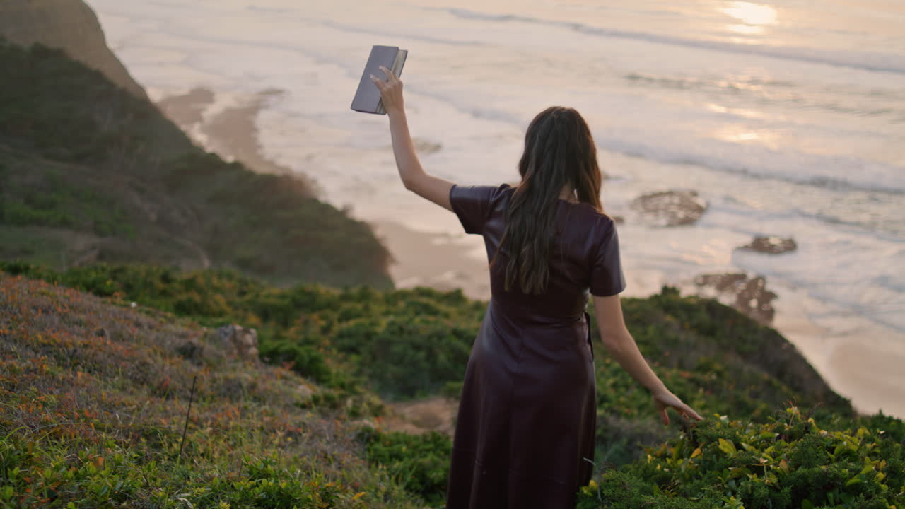 chica bajando de la costa al océano con un libro en la mano. vista trasera chica desconocida caminando por la colina