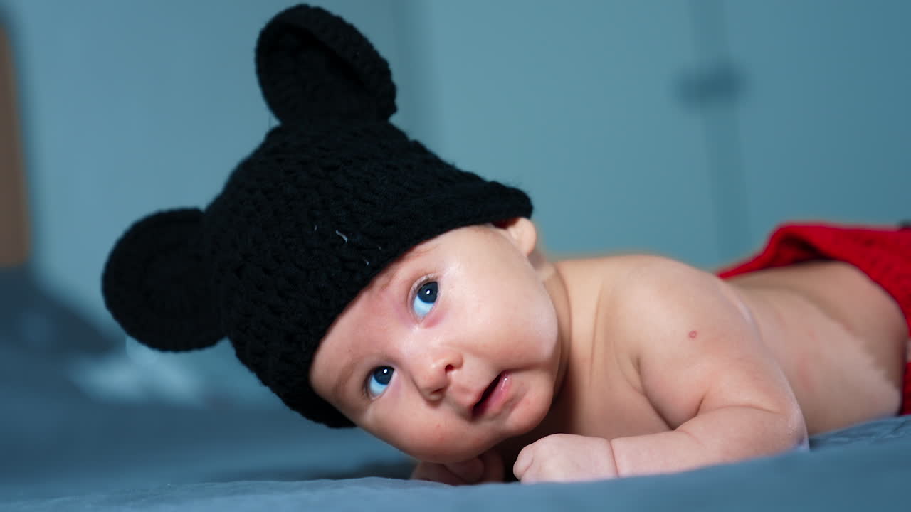 Lovely sweet baby in Mickey Mouse hat rises head while lying on the bed. Beautiful blue-eyed child looks up and opens tiny mouth. Close up.