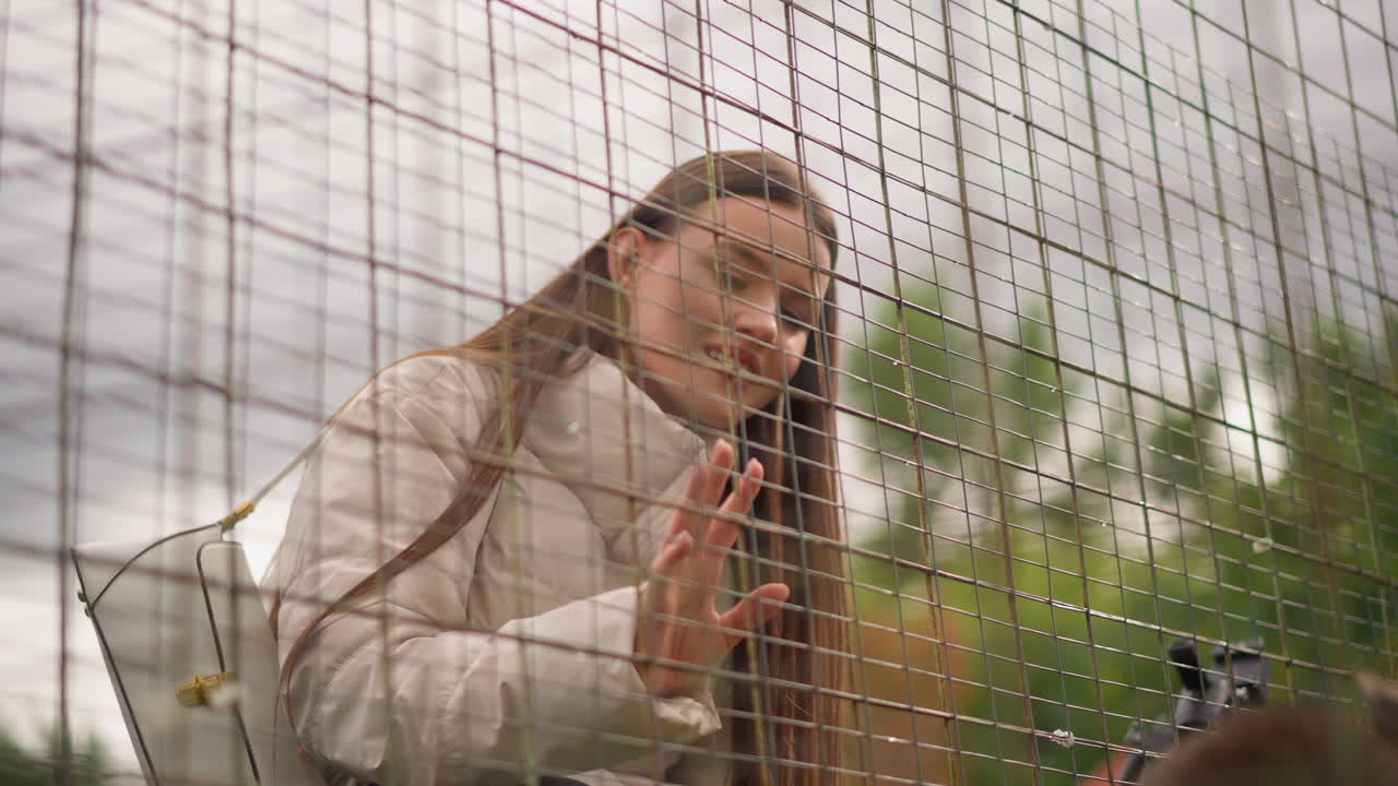 Donna al paddock che tocca la rete, interazione delicata con il cavallo attraverso la maglia, cielo nuvoloso e manto morbido, borsa a tracolla visibile, espressione contemplativa, messa a fuoco superficiale, sfondo naturale con vegetazione