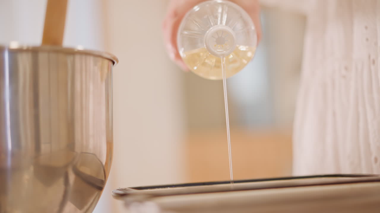 Mani che versano un filo d'olio nella padella prima di riempirla, bottiglia di plastica trasparente che rilascia un sottile flusso dorato, ciotola per mescolare e cucchiaio di legno sullo sfondo, versamento attento e misurato, calda preparazione in cucina per la cottura.