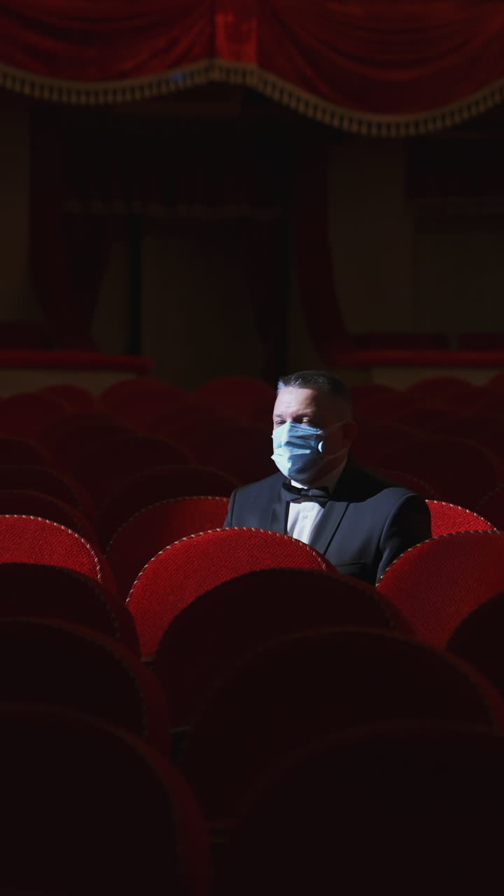 Rich man among rows of empty red chairs. Businessman in medical mask sitting in comfortable chairs alone on dark theater background. Pandemic in the world. Vertical video
