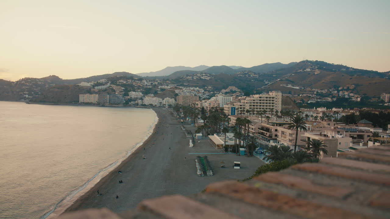 Campo lungo che mostra una tranquilla spiaggia fiancheggiata da palme ed edifici, con dolci colline sullo sfondo. Una tenue luce dorata aggiunge calma e profondità.
