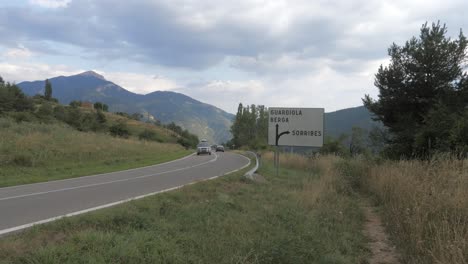 Cars-crossing-a-coutryside-road-with-moutains-in-the-background-on-a-cloudy-day,-passing-by-a-road-sign