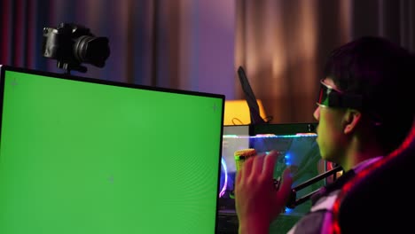 close up back view of asian boy streamer playing game, touching, and swiping experience virtual reality in futuristic goggles. green screen personal computer on desk illuminated by rgb led strip light