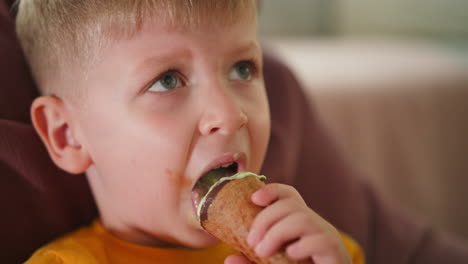 Close up of young boy with messy mouth enjoying ice cream cone while looking upward thoughtfully, wearing bright yellow shirt, seated comfortably on soft pink beanbag