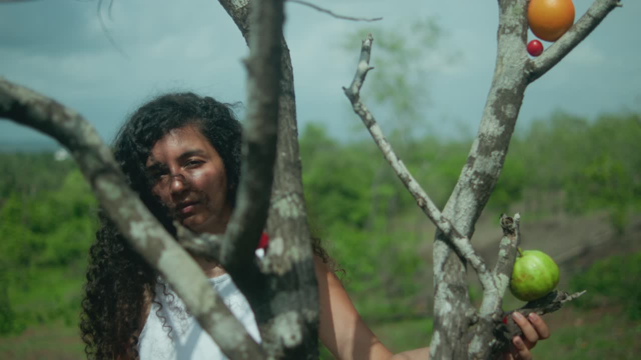 Pullback shot of a woman with long brown hair behind tree branches holding them, with fruits wedged between branches; green vegetation and blue sky in the background