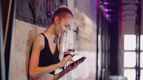 Mujer-Deportiva-Mensajería-De-Texto-A-Través-Del-Teléfono-Móvil-Mientras-Se-Apoya-En-La-Pared-Durante-El-Descanso-En-El-Gimnasio