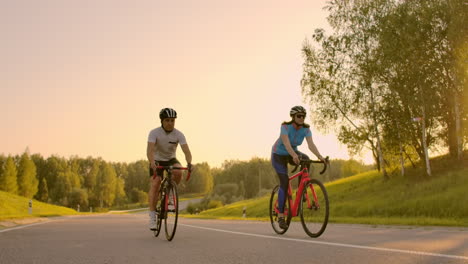 two cyclists a man and a woman ride on the highway on road bikes wearing helmets and sportswear at sunset in slow motion