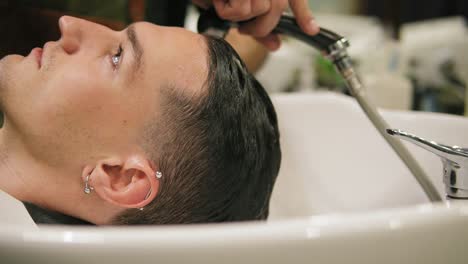 vista lateral de un joven elegante con un piercing en la oreja lavándose el cabello y un barbero irreconocible en un tiro de barbero