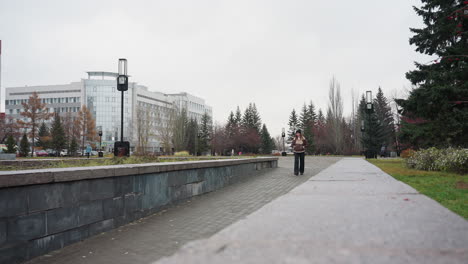 lady in black cap brown jacket walking slowly hands in pockets on stone pathway through urban park lined with pine trees modern buildings cloudy sky autumn atmosphere calm peaceful environment