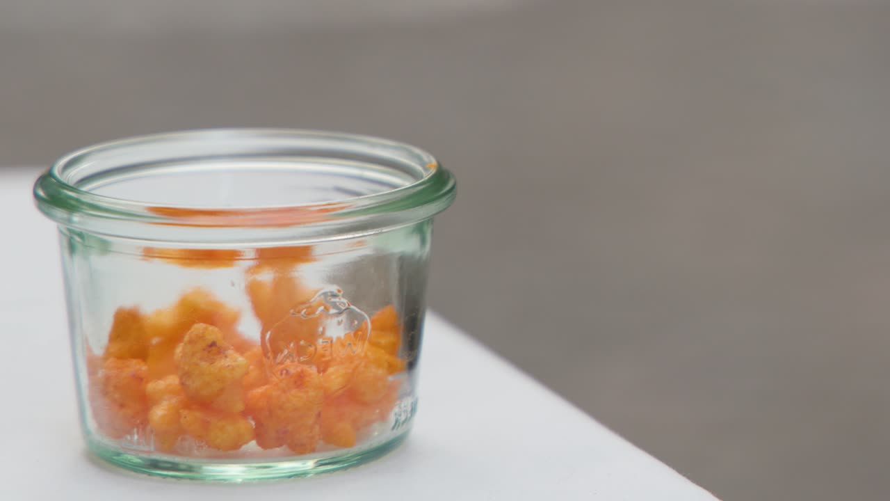 Hand picking orange popcorn from clear glass jar on white surface under natural lighting
