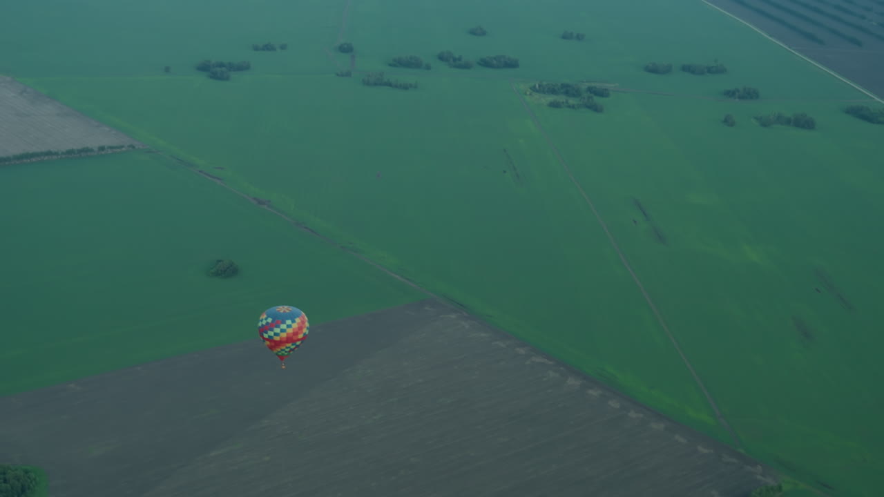 Close up of person filming vast green farmland from aerial perch, capturing distant colorful hot air balloon drifting over patchwork fields beneath soft pastel sky during golden hour flight