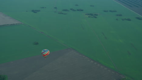 close up of person filming vast green farmland from aerial perch, capturing distant colorful hot air balloon drifting over patchwork fields beneath soft pastel sky during golden hour flight