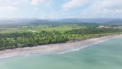 Aerial-view-of-empty-beach,-clear-Caribbean-Sea,-Palm-Tree-Plantation-and-mountains-in-Background---Miches,-Dominican-Republic-in-summer