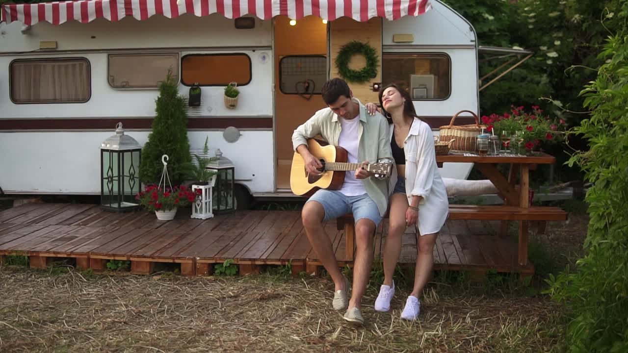 행복 하고 편안 한 부부 가 집 트레일러 앞 에서 야외 에 앉아 있고 남자 는 여자친구 를 위해 기타 를 연주 하고 있다. 야외 에서. 여자 는 음악 을 듣고 남자 를 포옹 하고 있다. 느린 동작. 초상