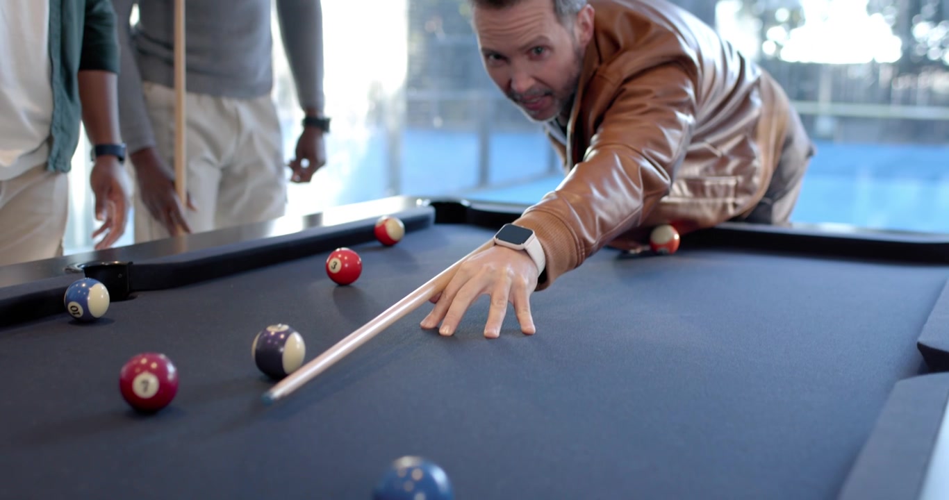Diverse men watching shooter lining up, hitting balls with cue during game on blue-felt table