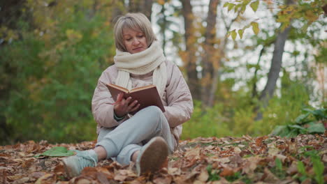 woman dressed in warm coat and scarf seated with crossed legs on forest ground focused on reading book, surrounded by dry autumn leaves and soft morning light filtering through colorful trees