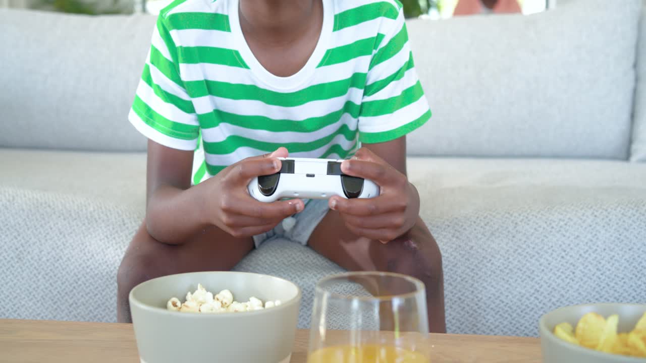 Upon game starting child holding pad pressing buttons grabbing popcorn and resuming play on sofa
