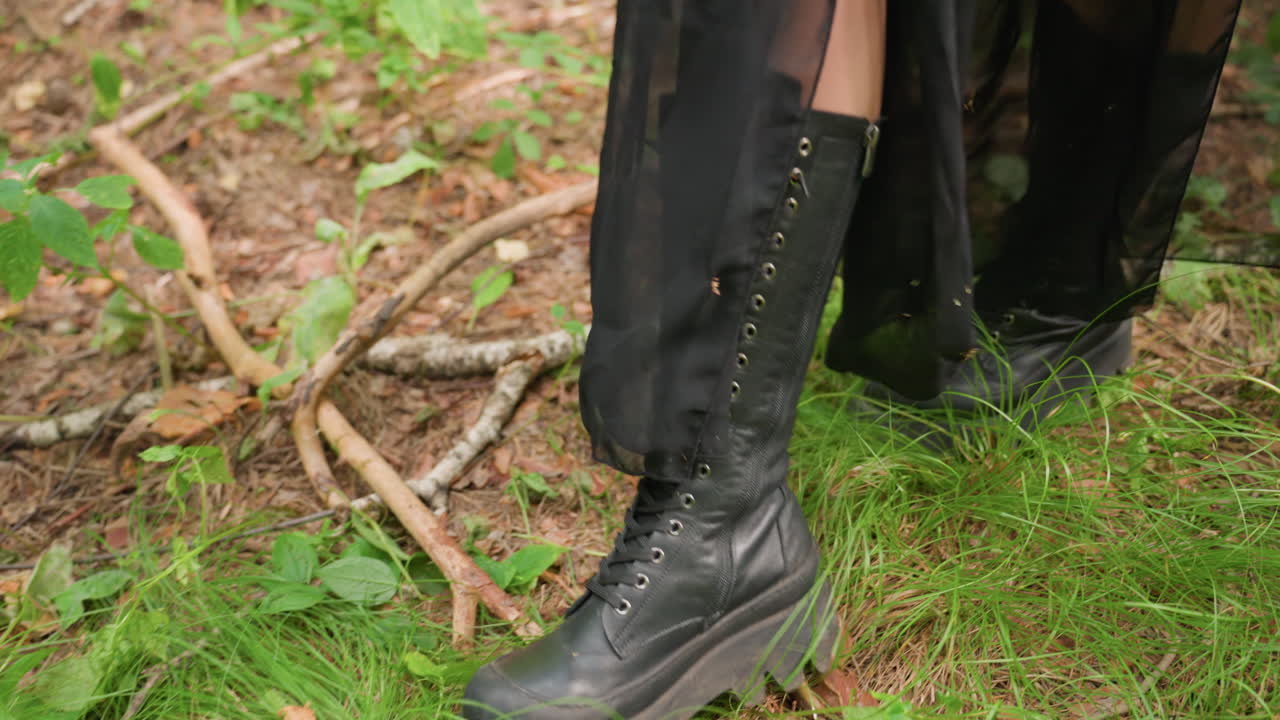 Close up of legs in black gown and boots stepping carefully through forest floor, maneuvering over tree roots and branches, fabric flowing with movement