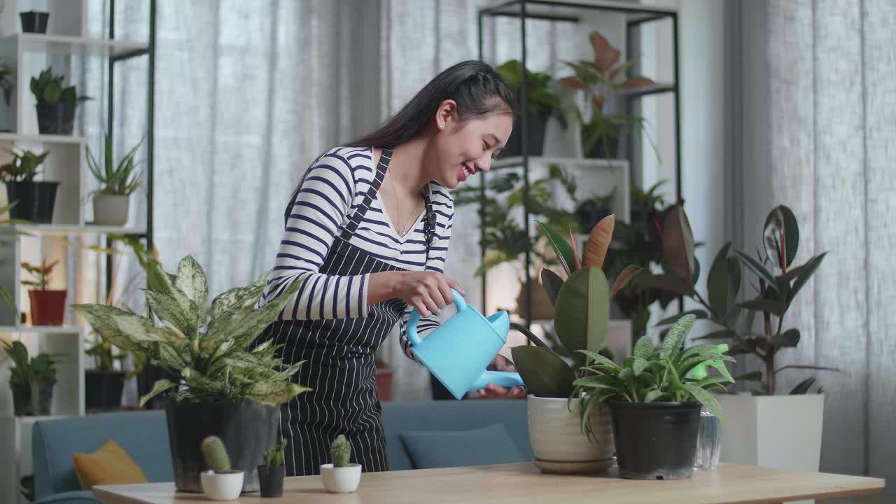 미소 짓는 아시아 여성 이 집 에 있는 식물 들 에게 물 을 주기 위해 물병 을 들고 있다