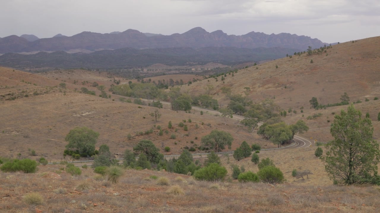 Veduta aerea di un'area dell'outback australiano, con una ripresa che svela le Flinders Ranges sopra dolci colline lungo una strada asfaltata, mostrando le montagne.