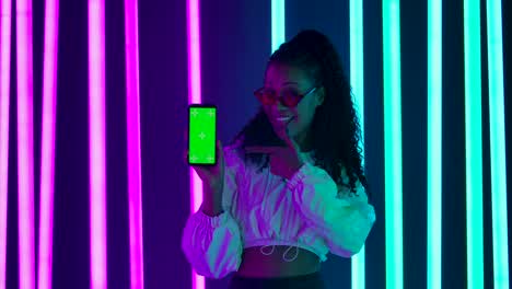 portrait of a young lovely female african american in stylish sunglasses points a finger at a smartphone with a green screen chroma key. slow motion