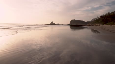 Playa-Maderas-beach-in-Nicaragua-with-shark-fin-rock-formation-far-away-during-sunset,-Handheld-ultra-wide-shot