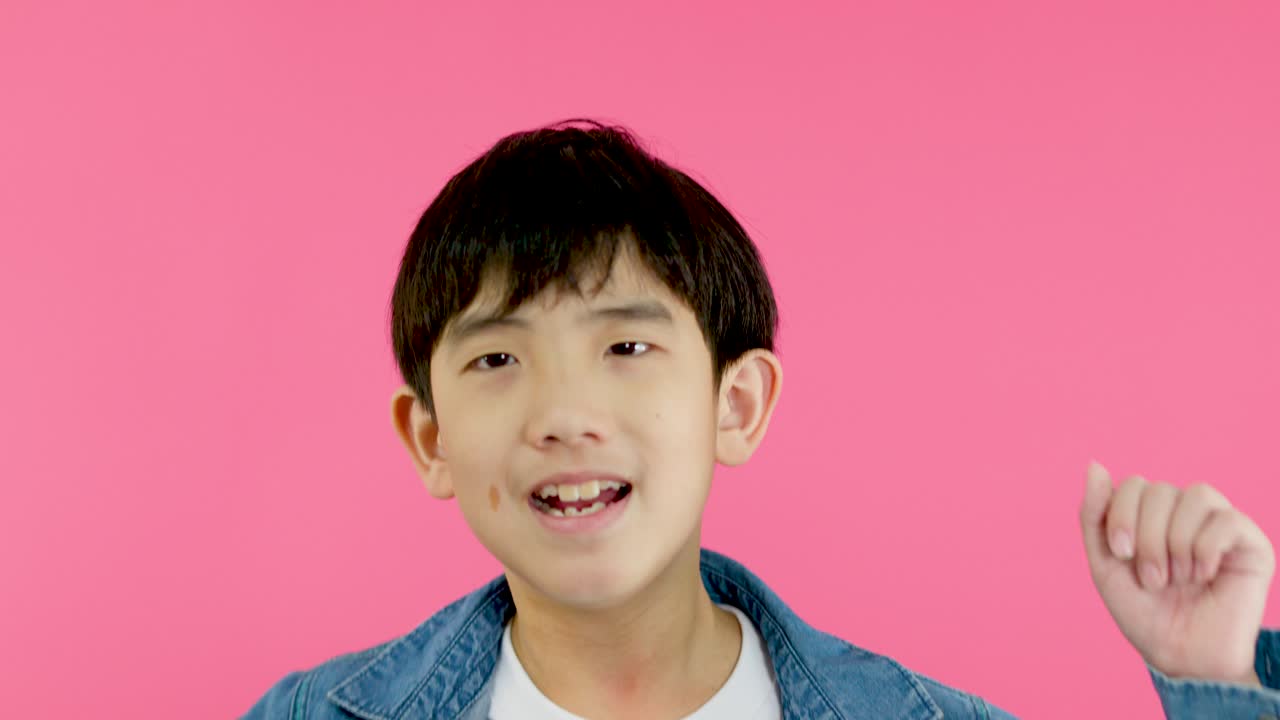 Young boy in denim jacket smiles, gestures OK hand sign, bright pink background, cheerful mood