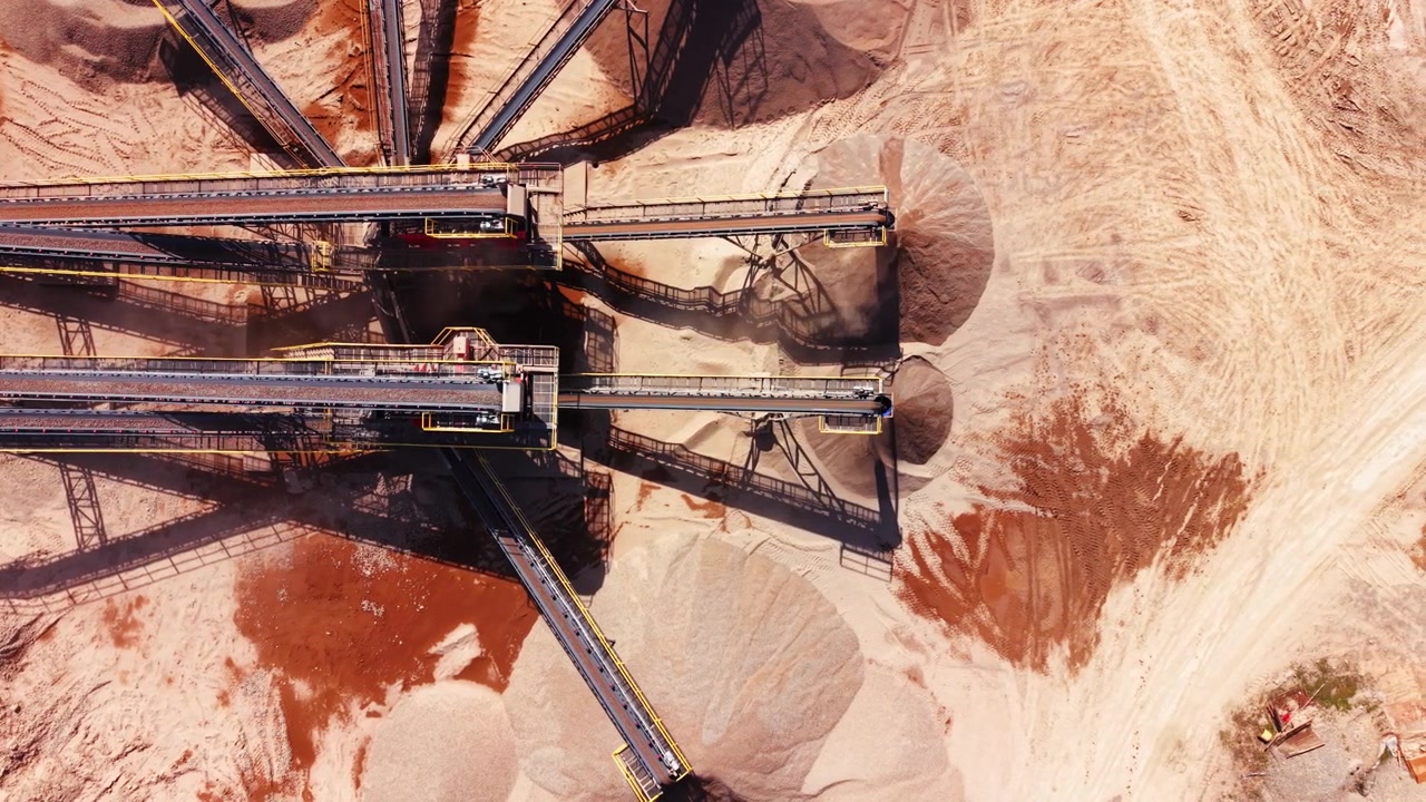 Double industrial belts transporting crushed rock to sand stockpiles below. Top-down aerial view of parallel aggregate conveyor belts.