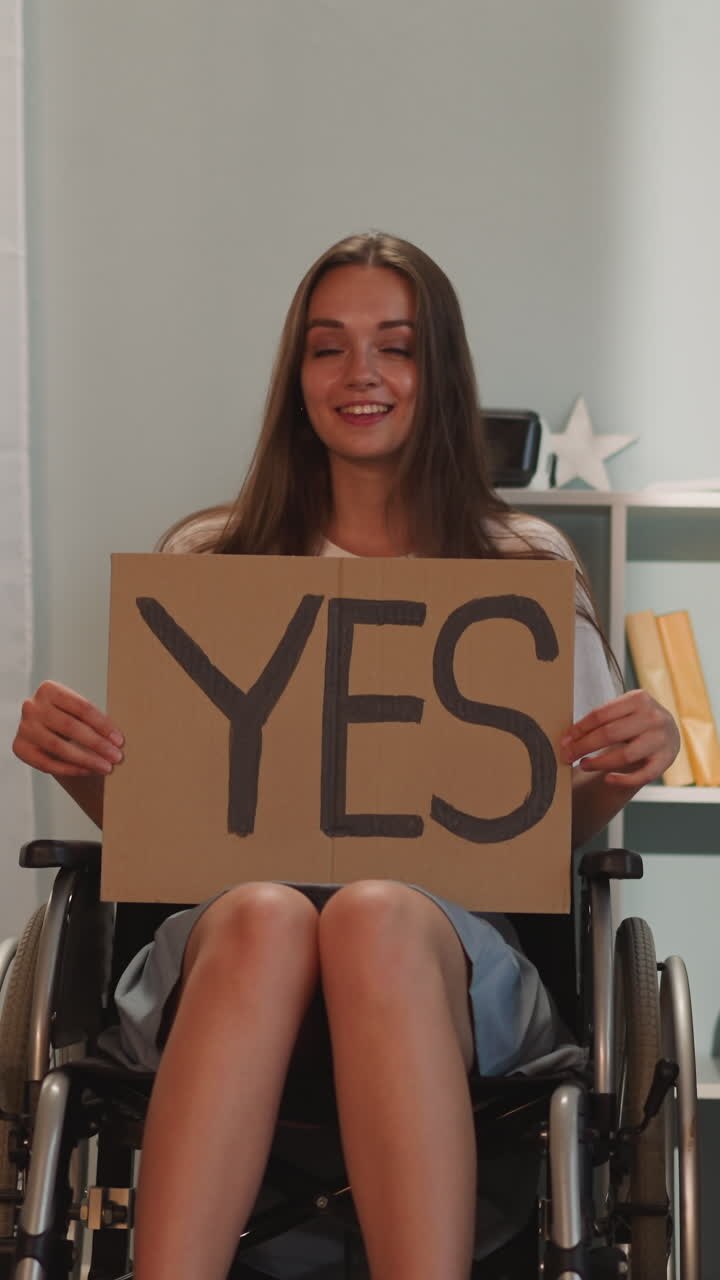 Positive brunette woman turns poster with word Yes to camera leaning head to side. Young person with disability sits in wheelchair smiling