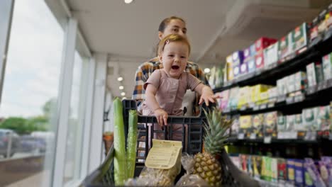 Porträt-Eines-Glücklichen-Kleinen-Mädchens,-Das-In-Einem-Einkaufswagen-Sitzt,-Während-Es-Mit-Seiner-Mutter-In-Einem-Supermarkt-Einkauft