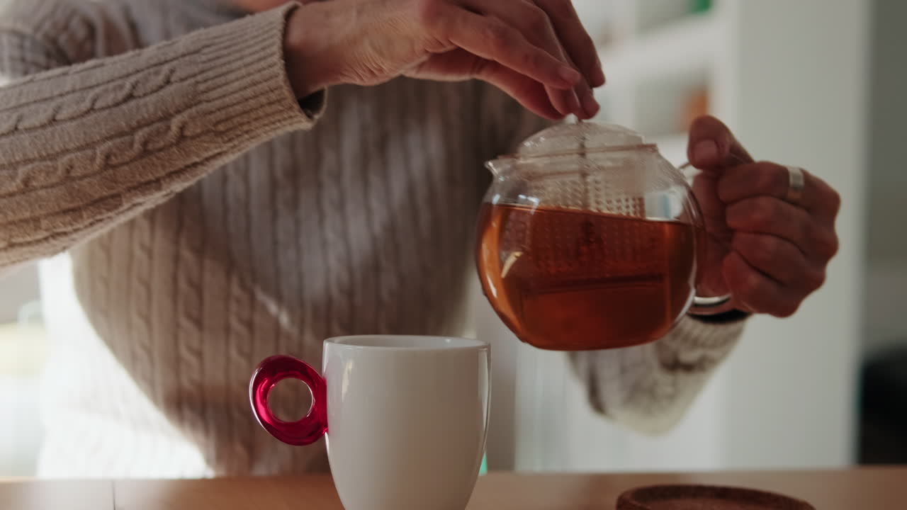 Close-up of a Person Pouring Hot Tea from a Glass Teapot into a White Mug